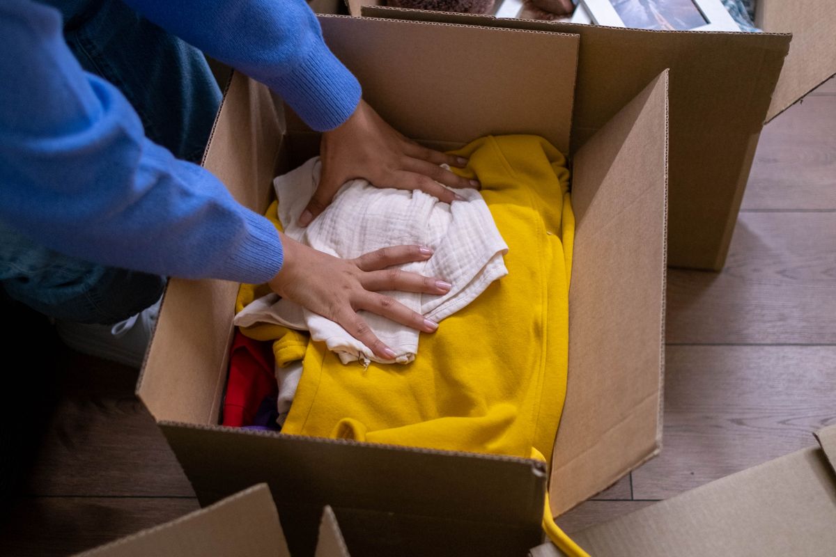 Hände legen Kleidungsstücke behutsam in einen Umzugskarton, daneben weitere Kartons mit Bilderrahmen